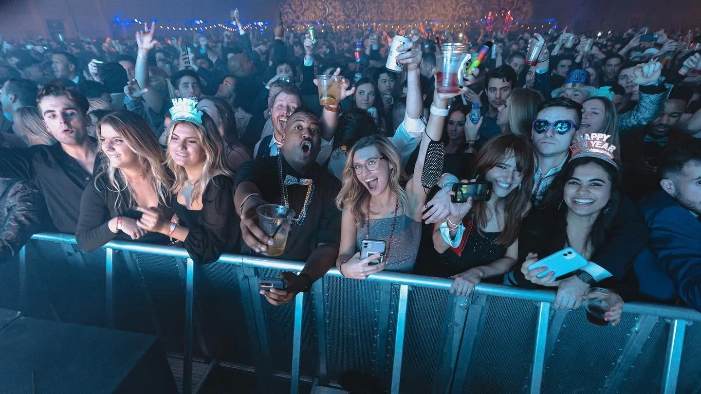 Big Night New Year’s Eve guests in tiaras and party hats cheer at the front-row barrier, drinks and phones raised