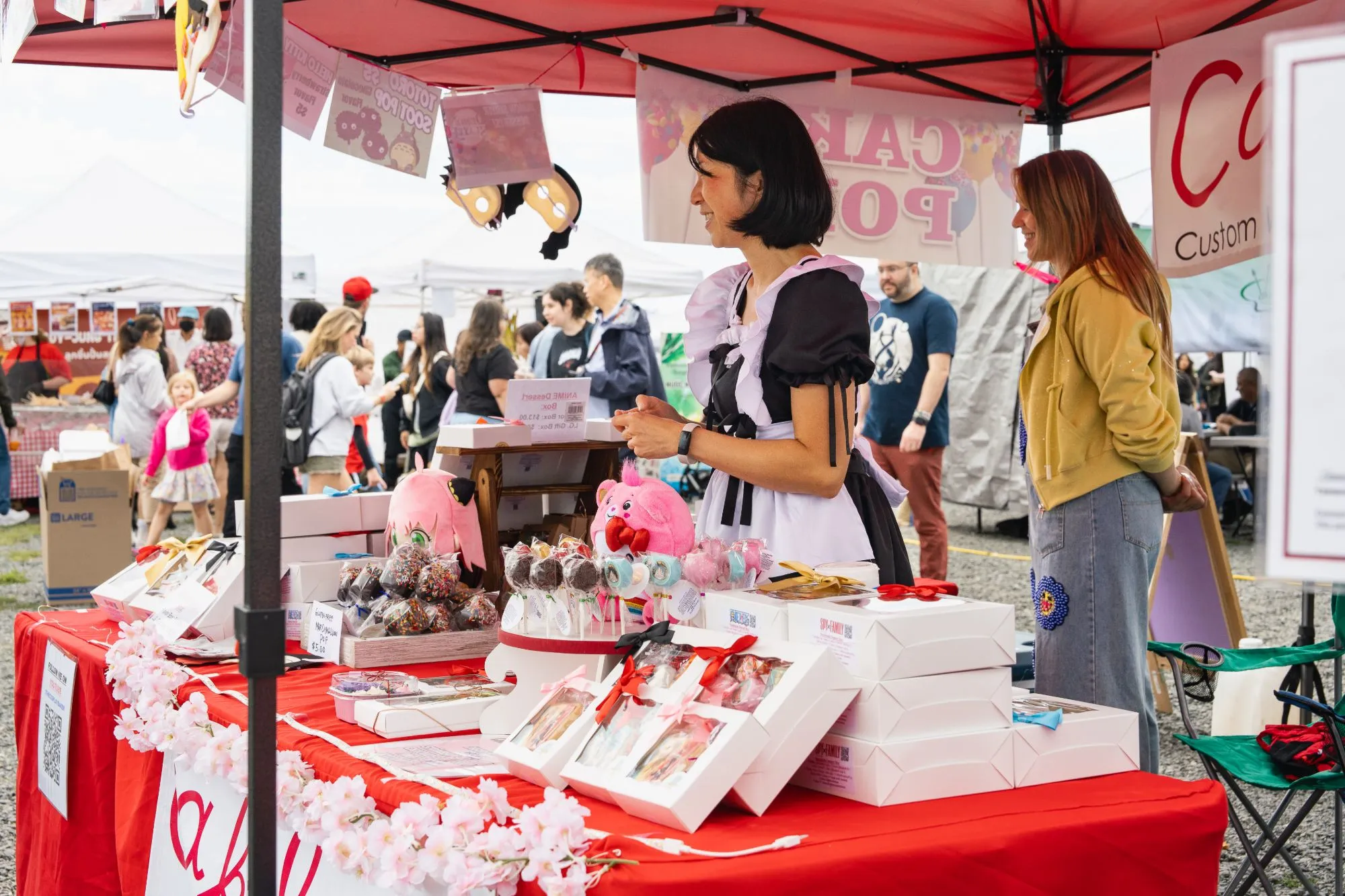 US Asian Fest vendor in a maid costume sells anime-themed cake pops and dessert boxes at a red-tented booth